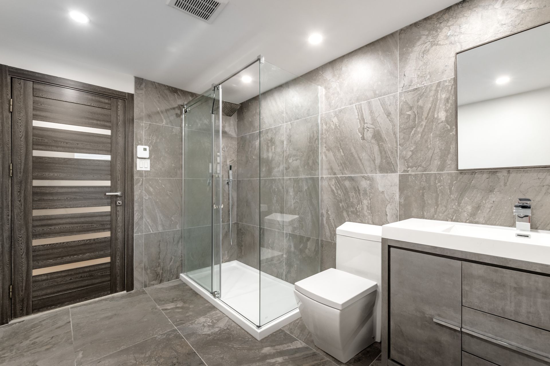 Modern gray-tiled bathroom with glass shower, vanity, toilet, and wood door