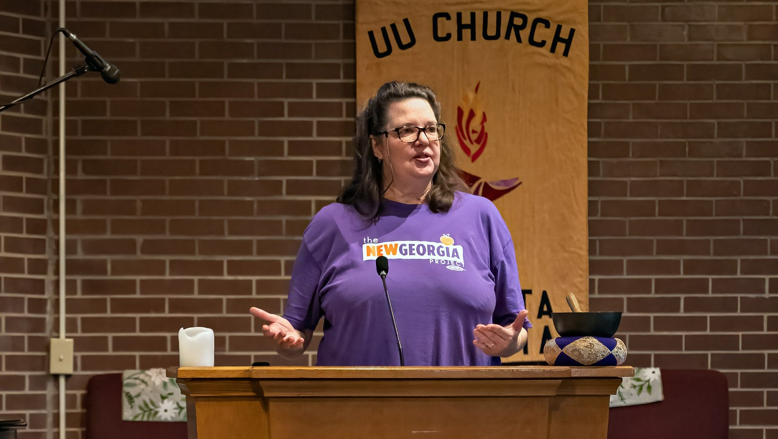 A woman in a purple shirt is standing at a podium in front of a microphone.