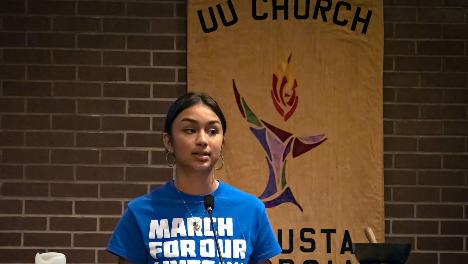 A woman in a blue shirt is standing in front of a microphone in front of a brick wall.