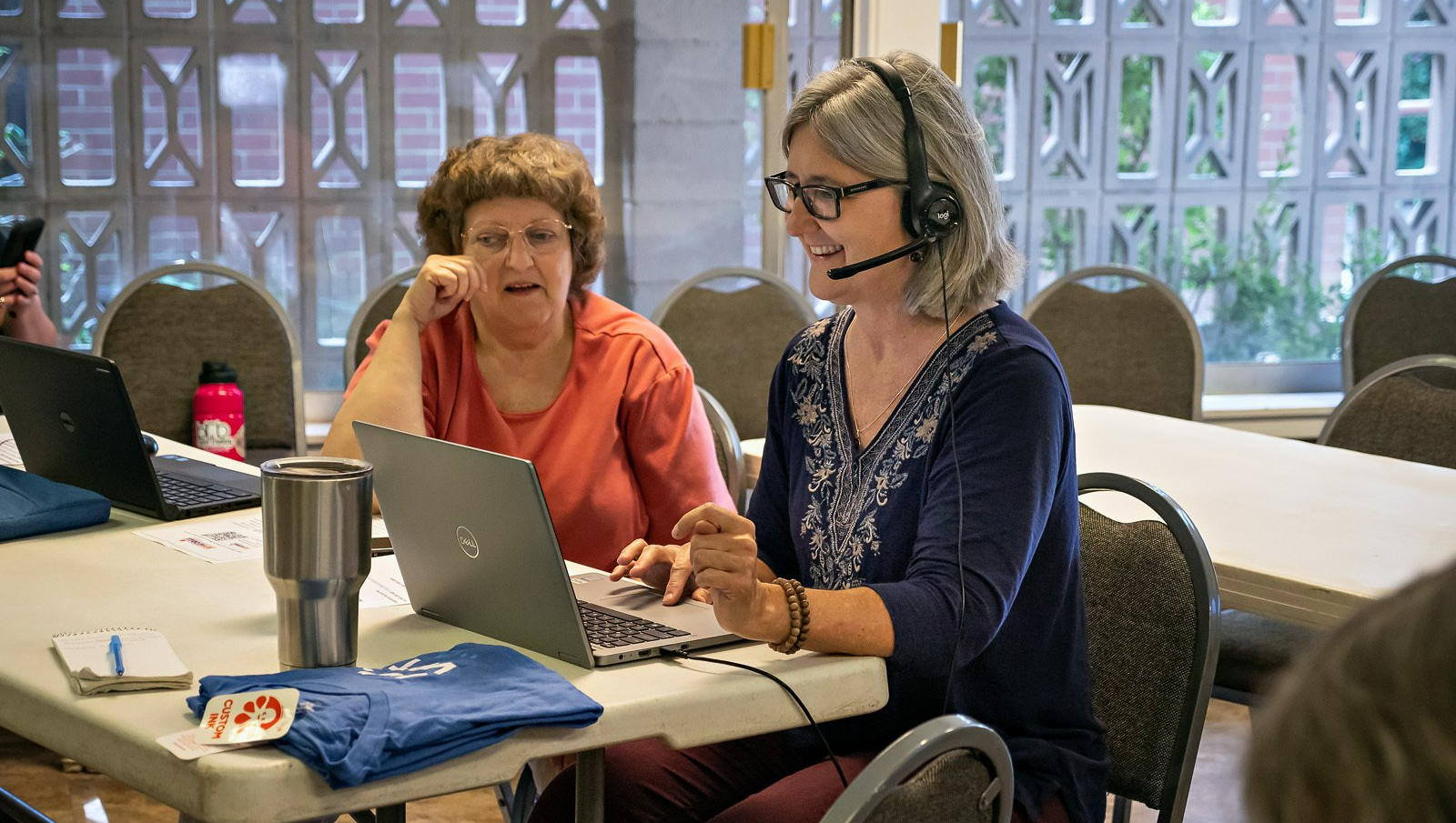 Two women are sitting at a table with laptops and headphones on.