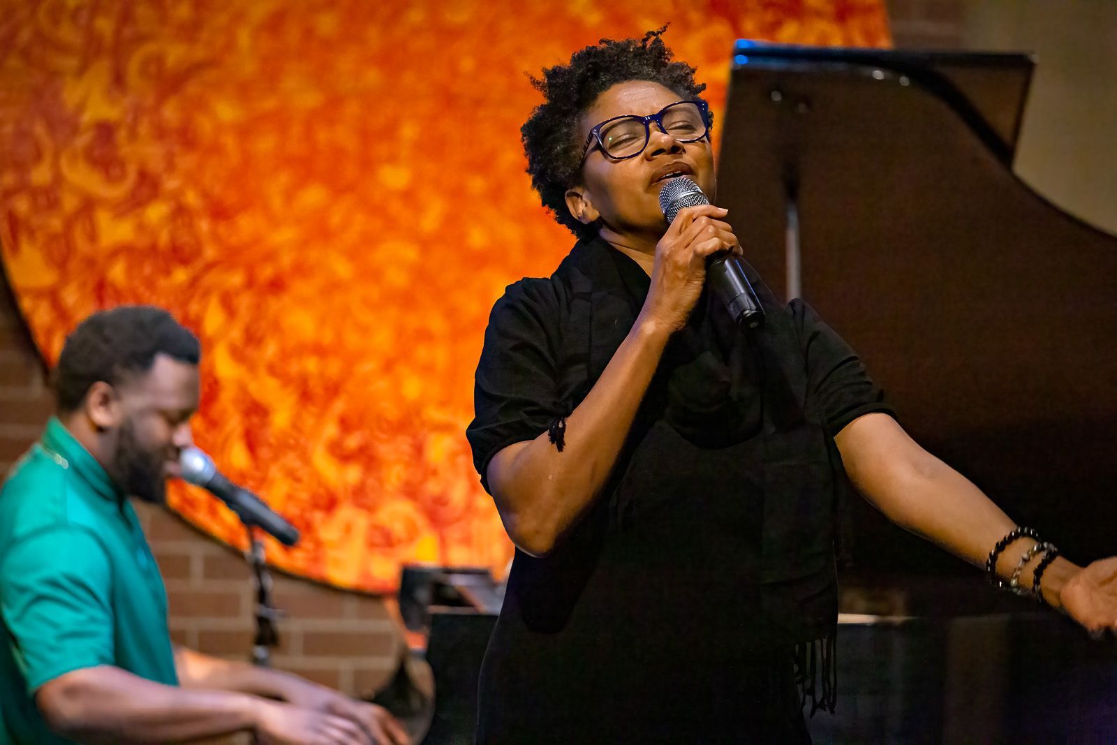 A woman is singing into a microphone while a man plays a piano.