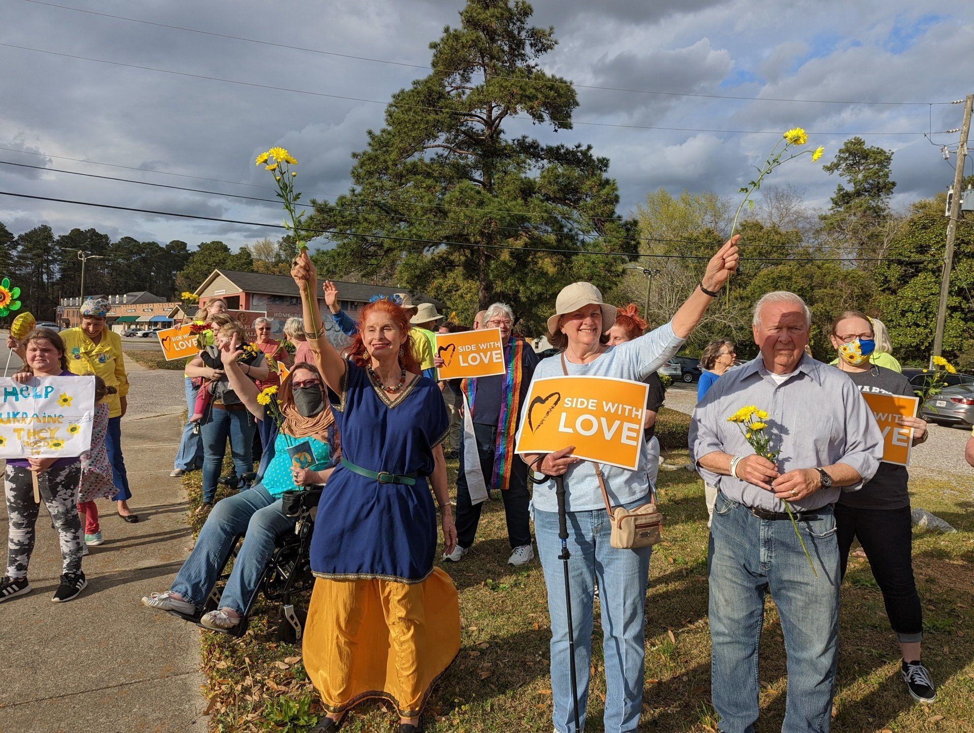 A group of people are standing by a road holding signs and flowers.
