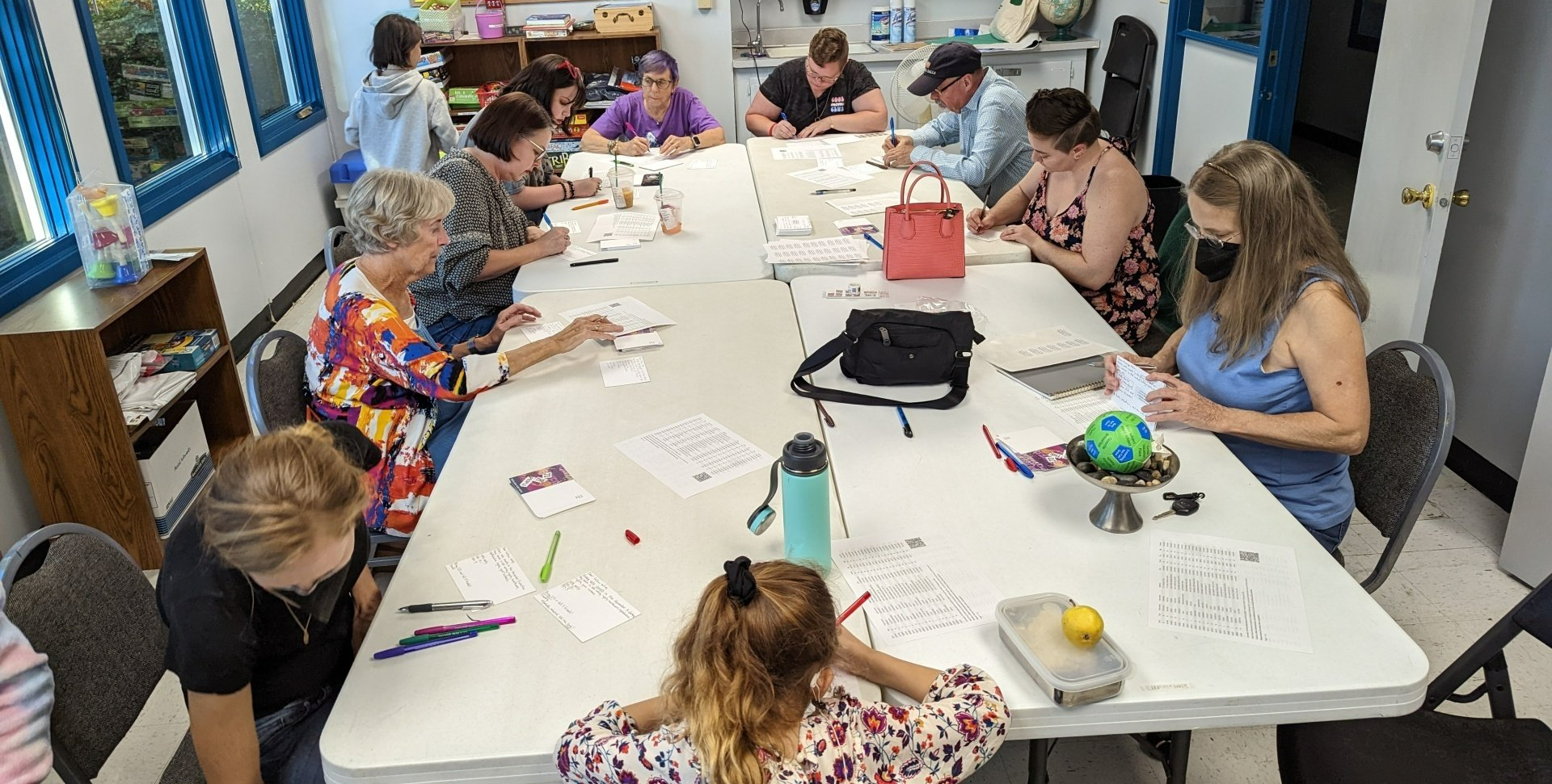 A group of people are sitting around a long table in a room, writing postcards.