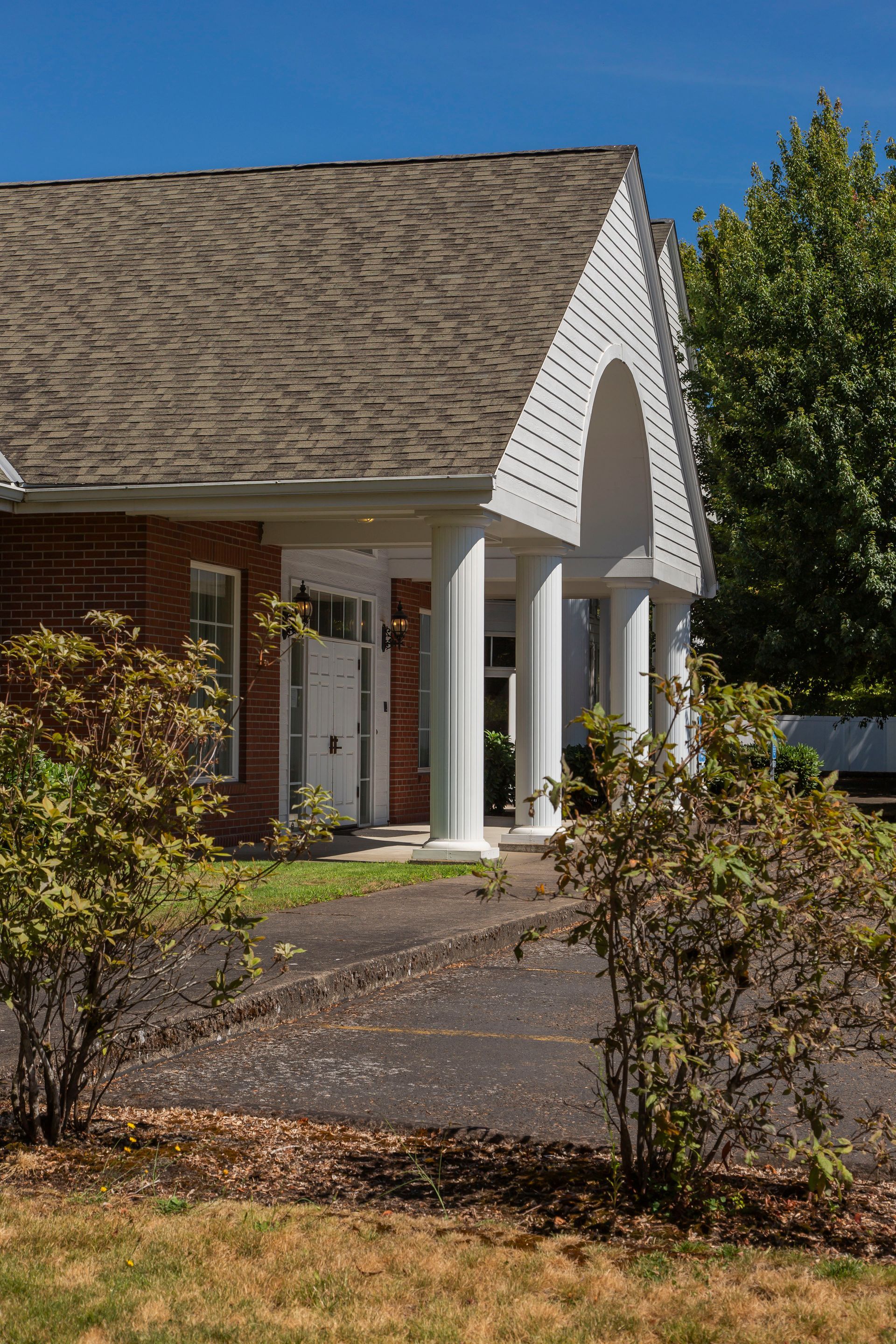 Brick building with white pillars and arched entryway, asphalt driveway, and shrubbery under a blue sky.