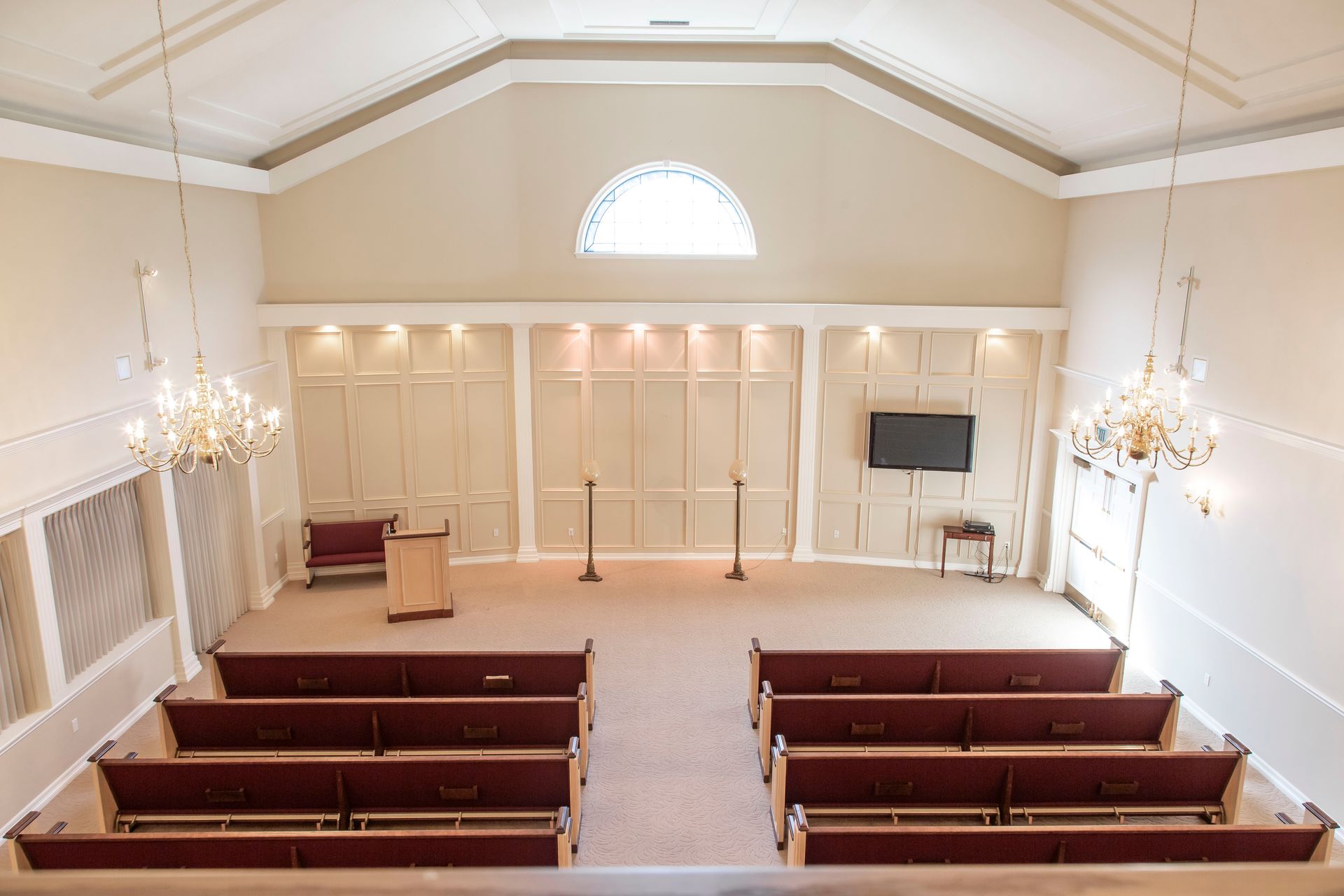 Interior of a chapel with rows of red pews, a podium, and chandeliers; light beige walls and carpet.