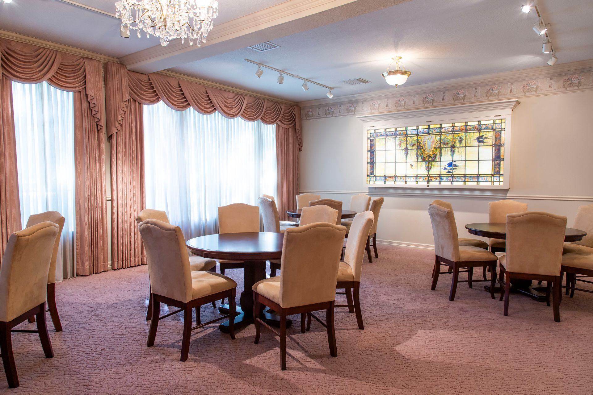 A room with round tables, chairs, pink carpet, curtains, and a chandelier.