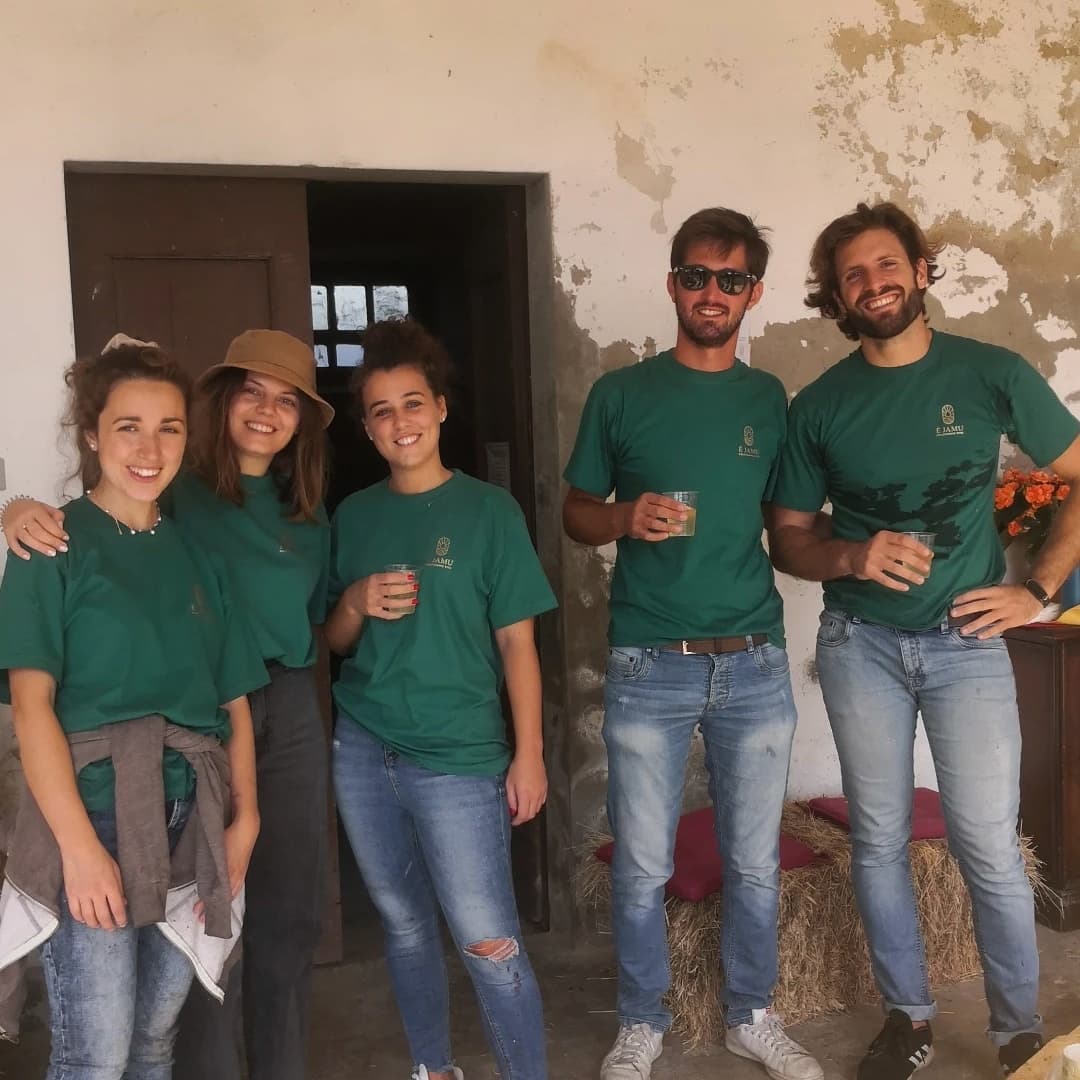 Five people in green shirts and jeans pose outdoors. One woman wears a hat. Some hold drinks.