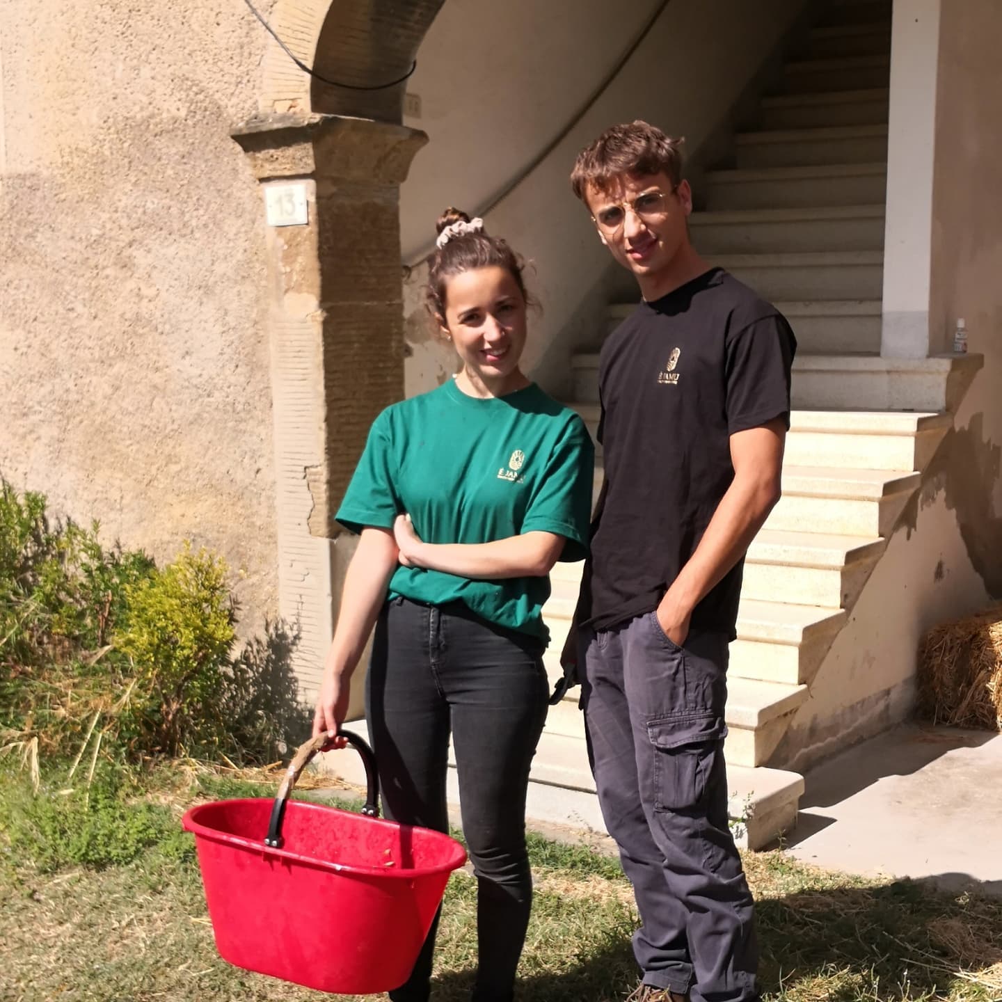 Winemakers of Ejamu standing by building; the woman holds a red bucket.