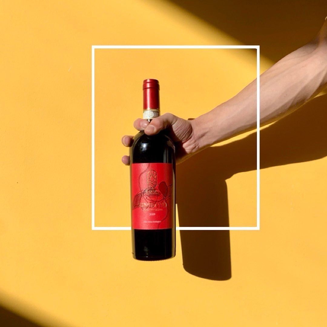 Hand holding a red Ejamu wine bottle with red label against a yellow wall
