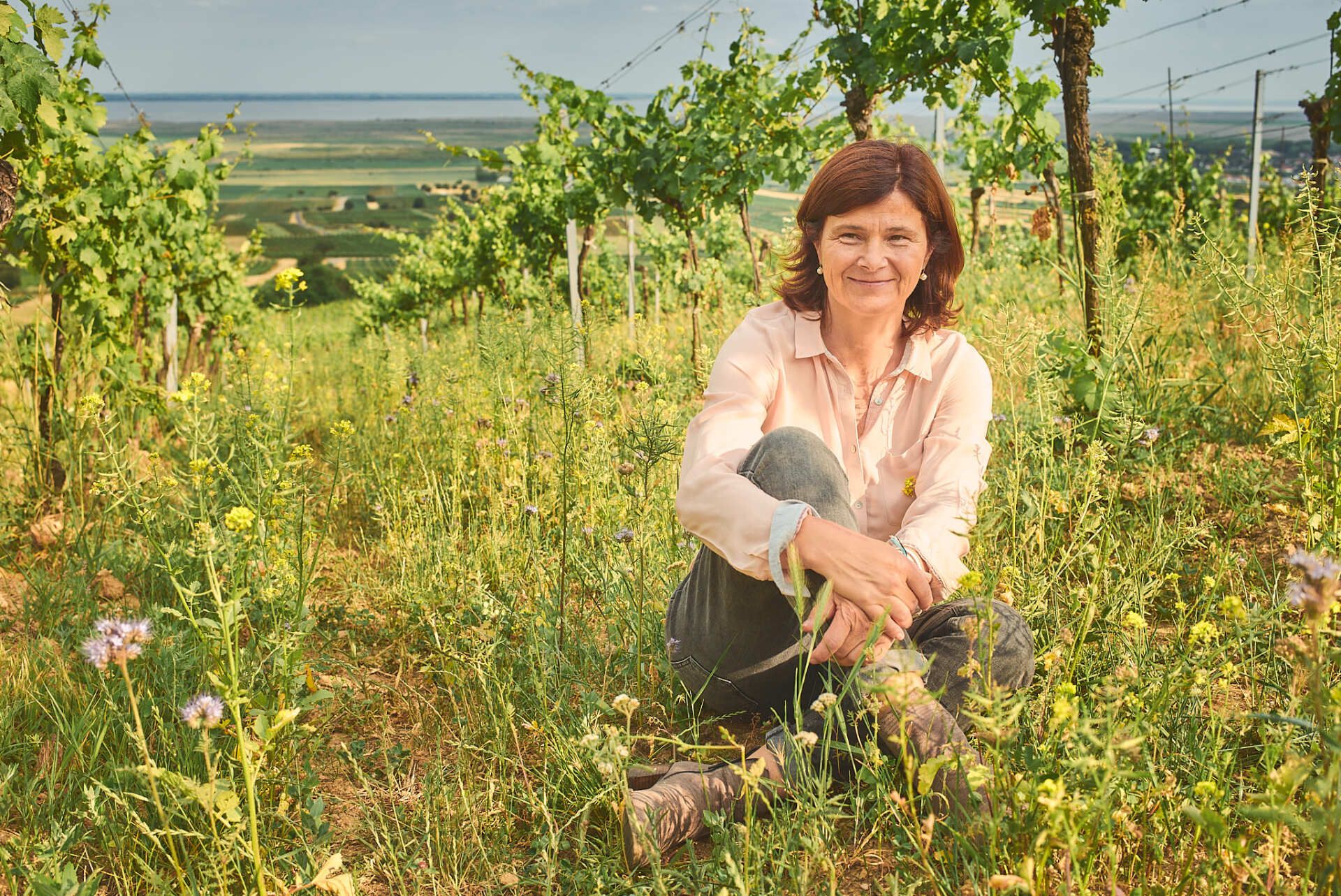 Birgit Braunstein sitting in a vineyard field with rows of vines, smiling, wearing a light pink shirt and jeans.