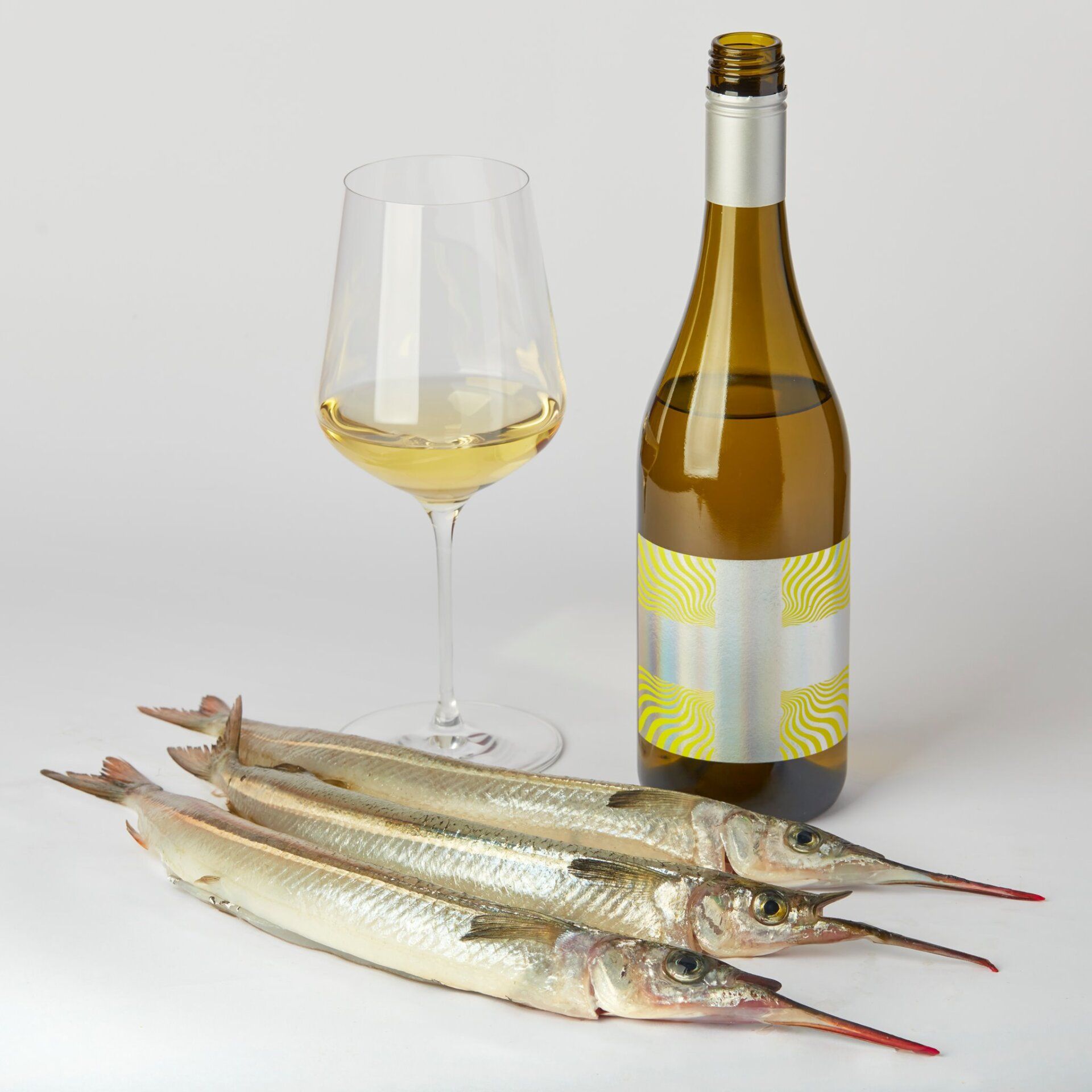 A glass of white wine, bottle of wine, and three needlefish on a white surface.