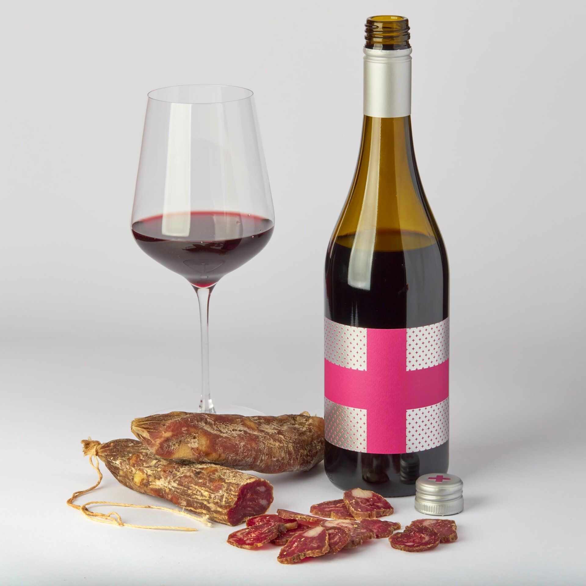 Bottle of red wine, glass of wine, and cured meat on white surface.