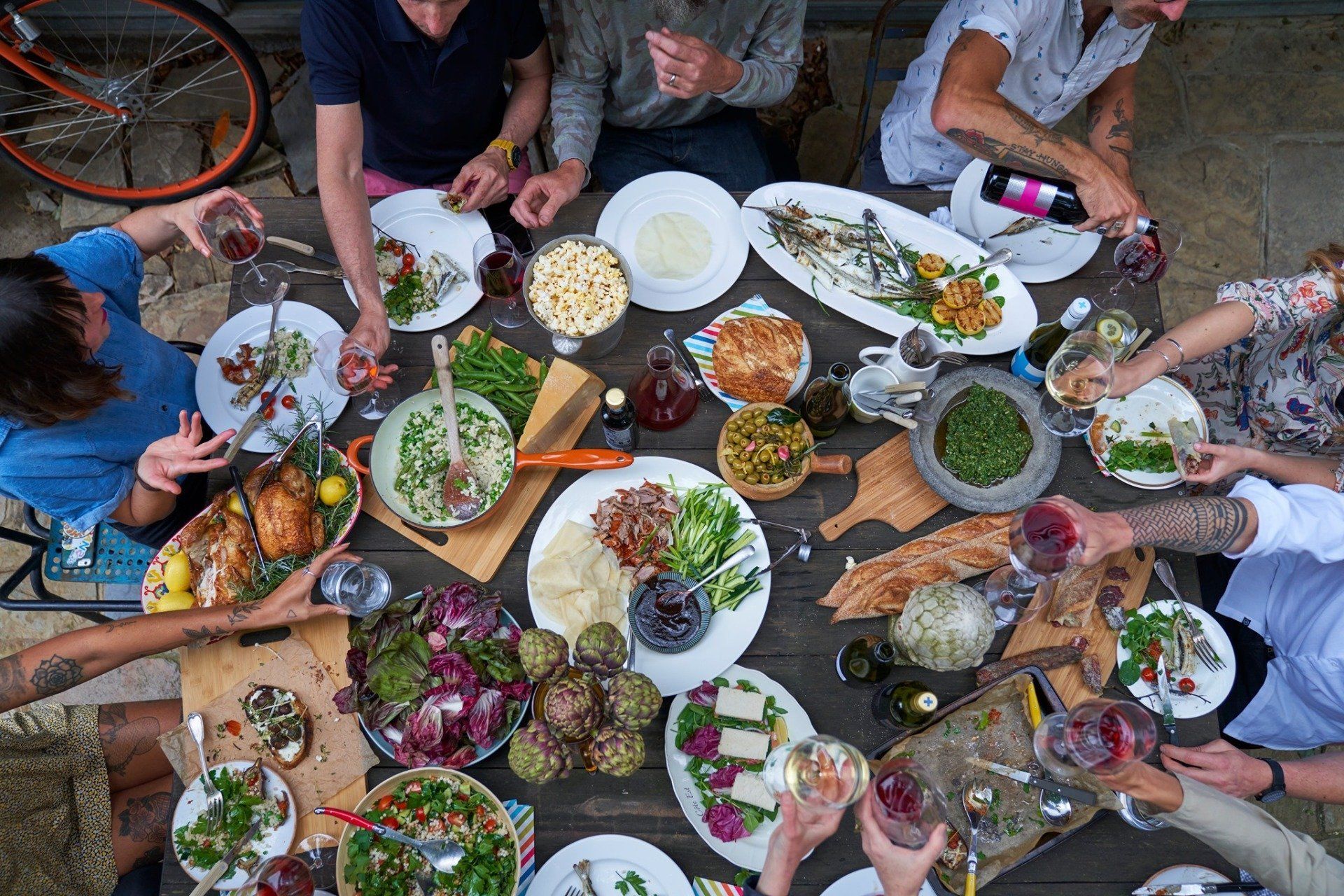 People around a table sharing a meal outdoors; various dishes and wine bottles present.