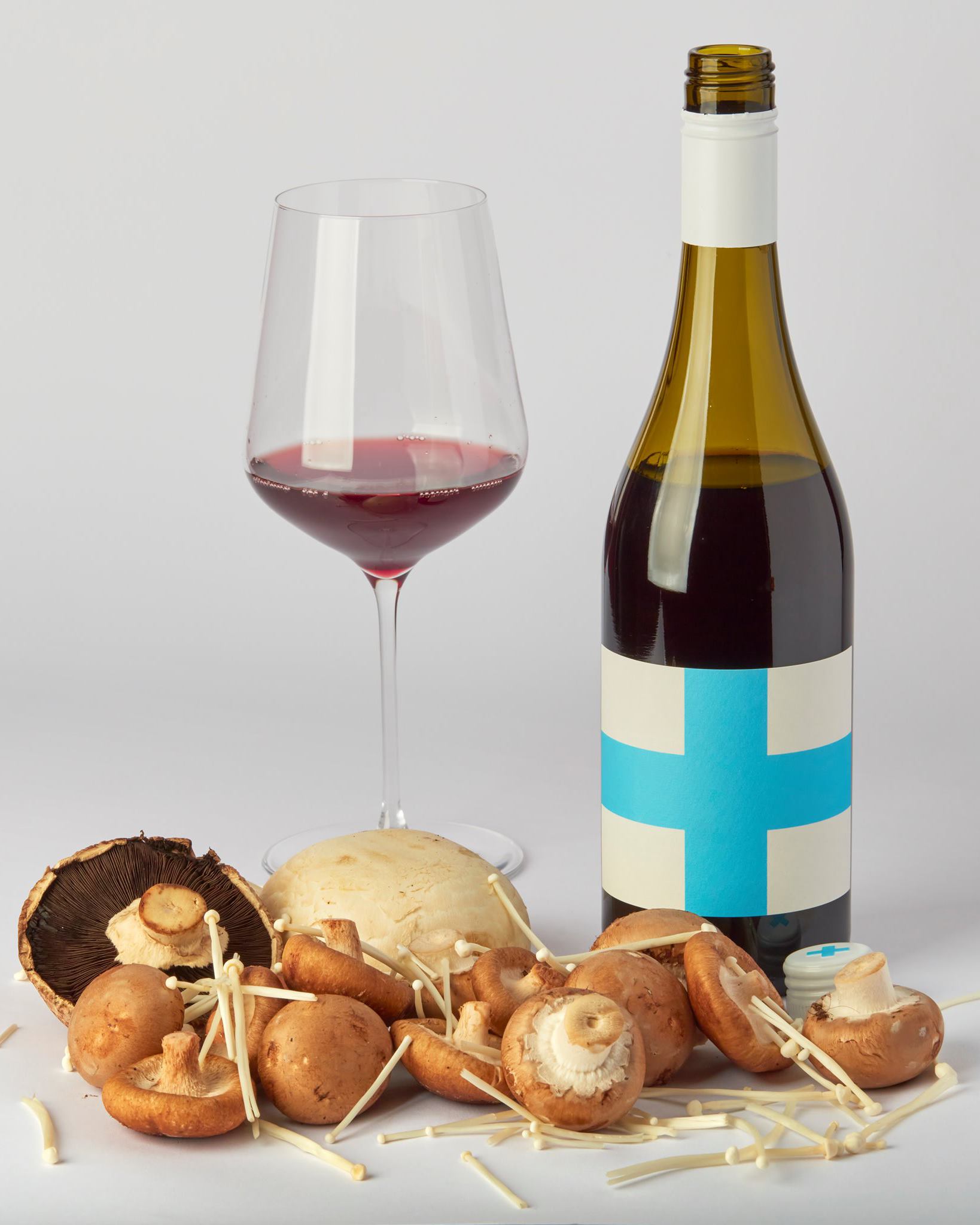 Bottle of red wine, glass of red wine, and mushrooms on a white surface.