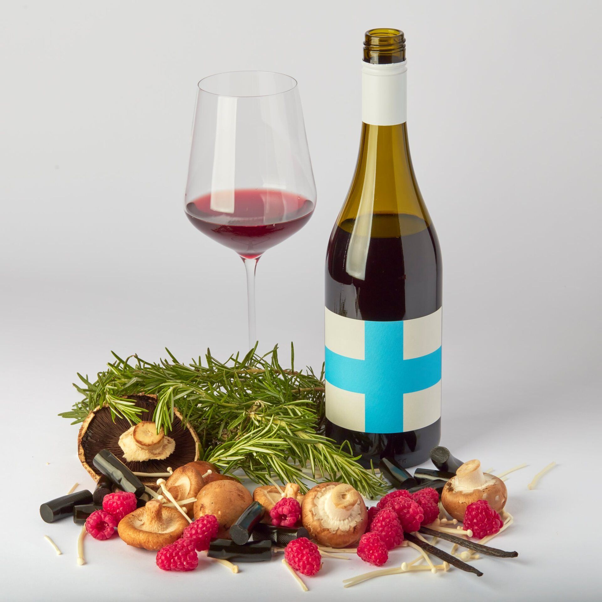 Wine bottle, glass of red wine, mushrooms, rosemary, raspberries, liquorice, and straw against white background.