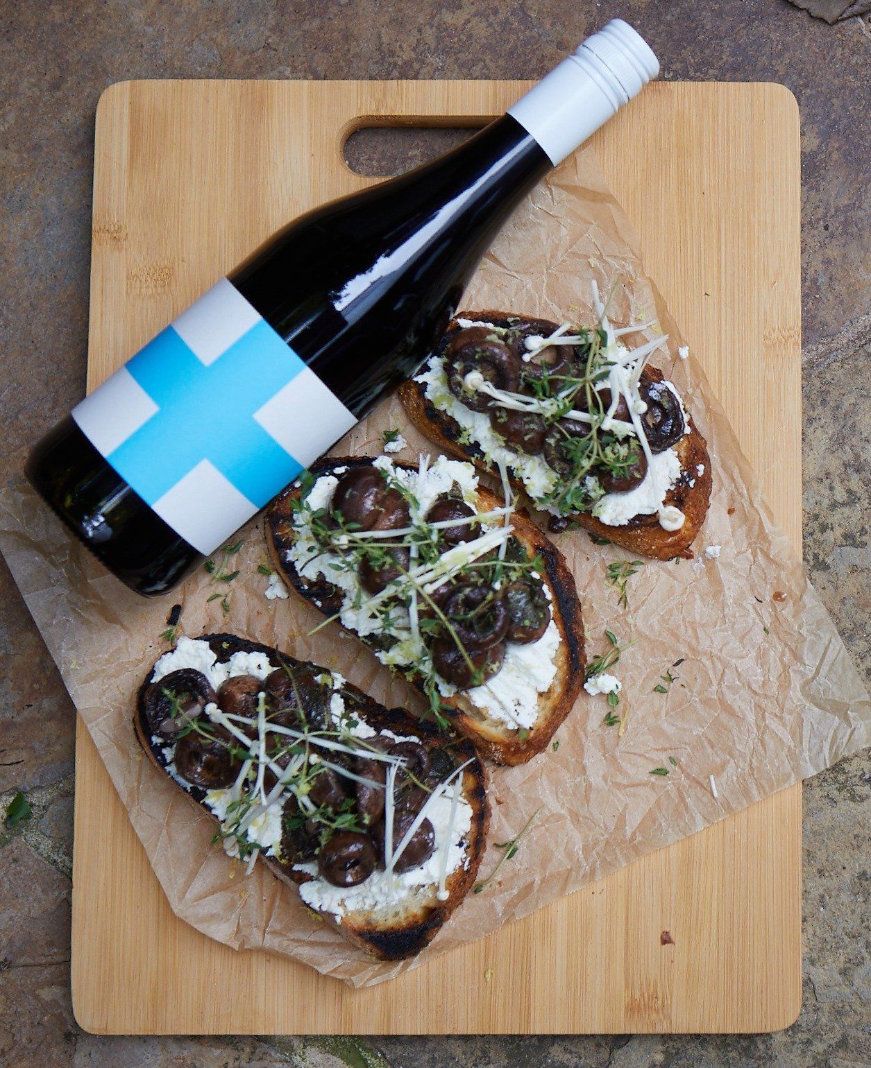 Bottle of wine next to toasted bread with olives, cheese, and herbs, on a wooden board.