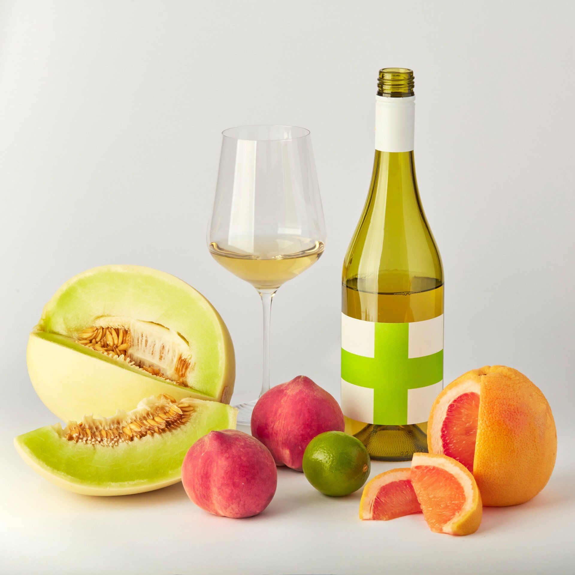 White wine bottle, glass, and fresh fruit: melon, peaches, lime, grapefruit, on white surface.