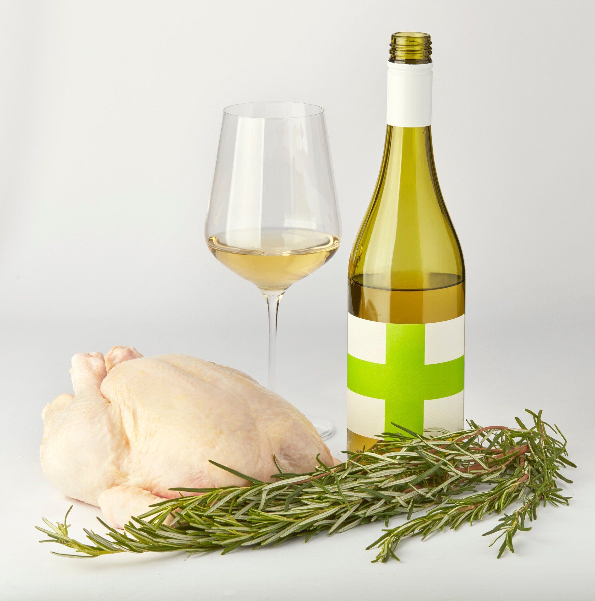 Raw chicken, white wine, rosemary, and a wine glass on a white surface.