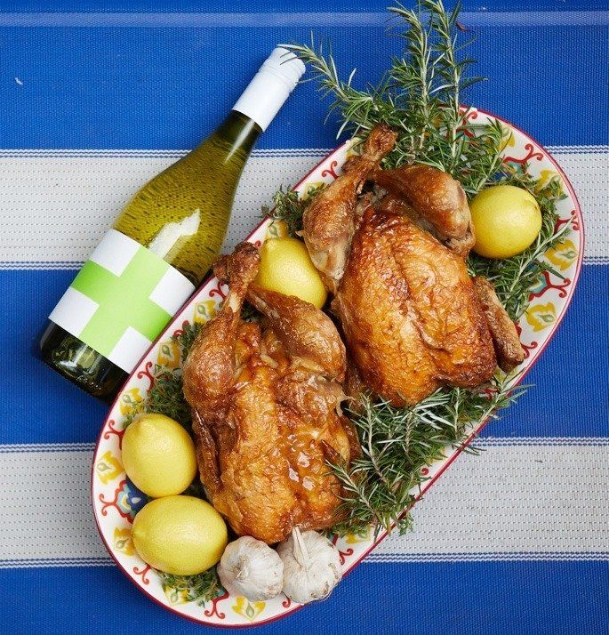Roasted chicken platter with lemons, rosemary, garlic, and a Save our Souls wine bottle on striped blue surface.