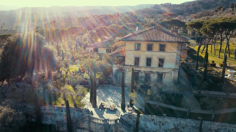 Sunlit villa perched on a hill, surrounded by trees and a stone wall.