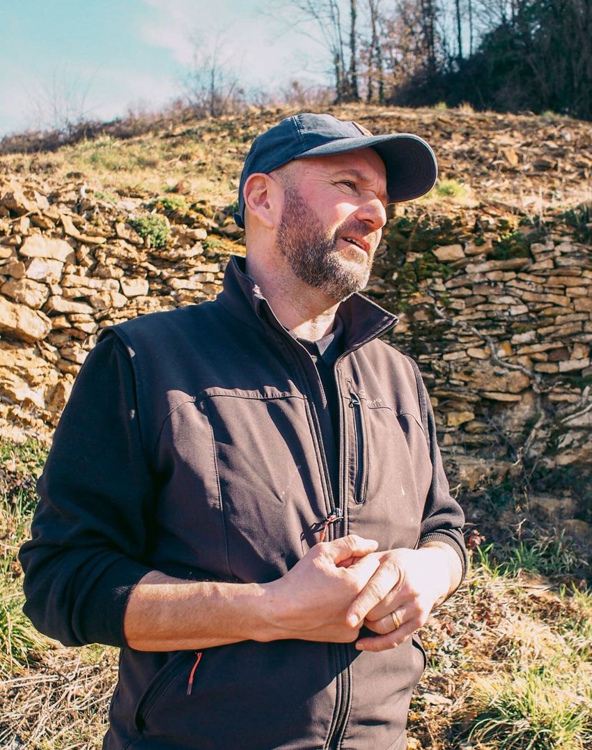 Man in a cap and vest, outdoors, looking to the side. Behind him is a stone wall and hillside.