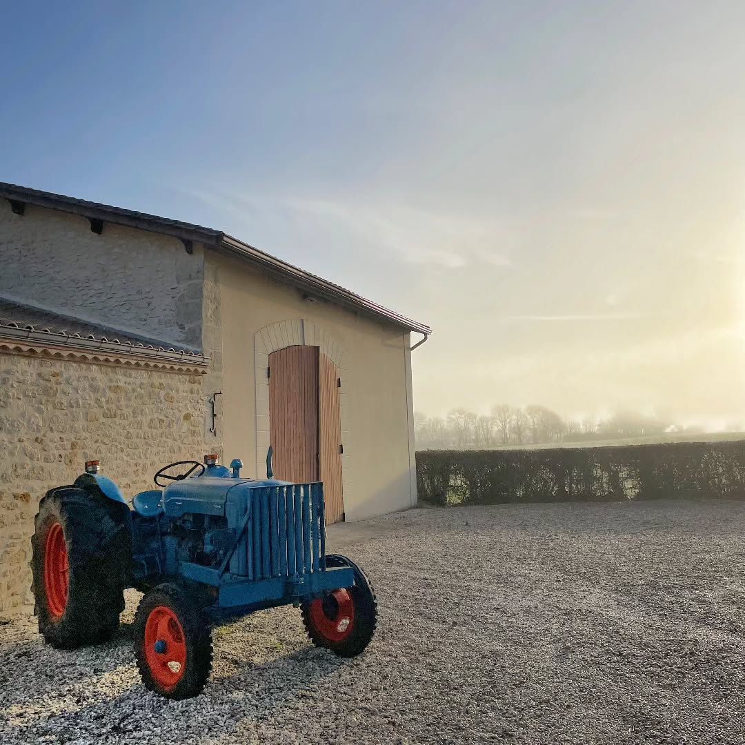Blue tractor with orange wheels parked by a stone building on a gravel lot, at Château Saint Hilaire