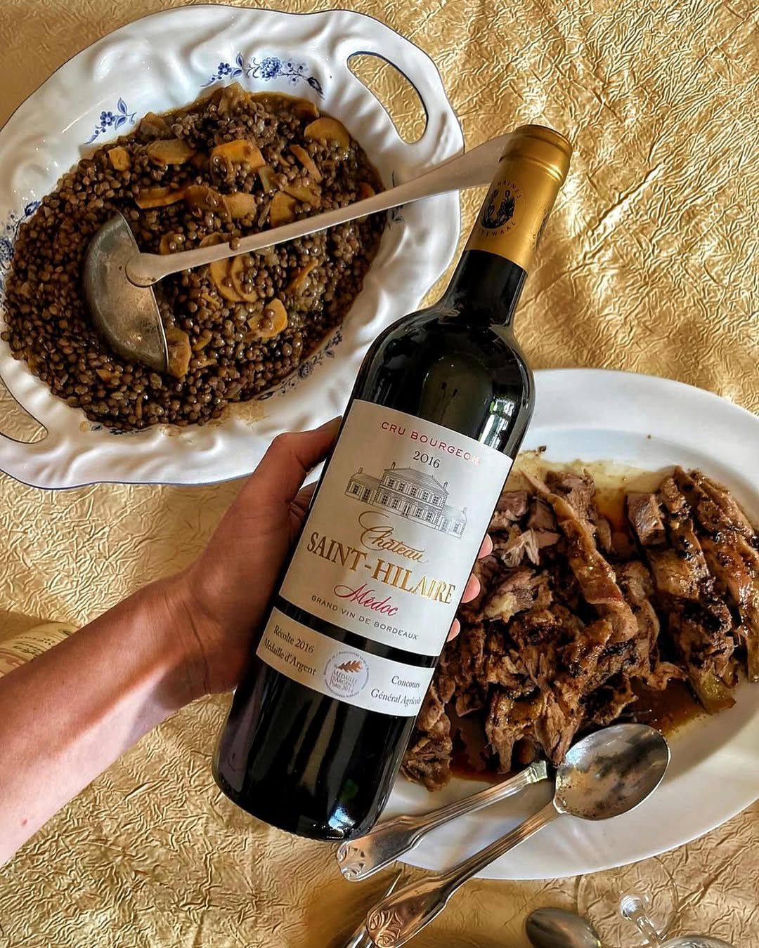 Hand holding a red wine bottle next to plates of food, at Château Saint Hilaire