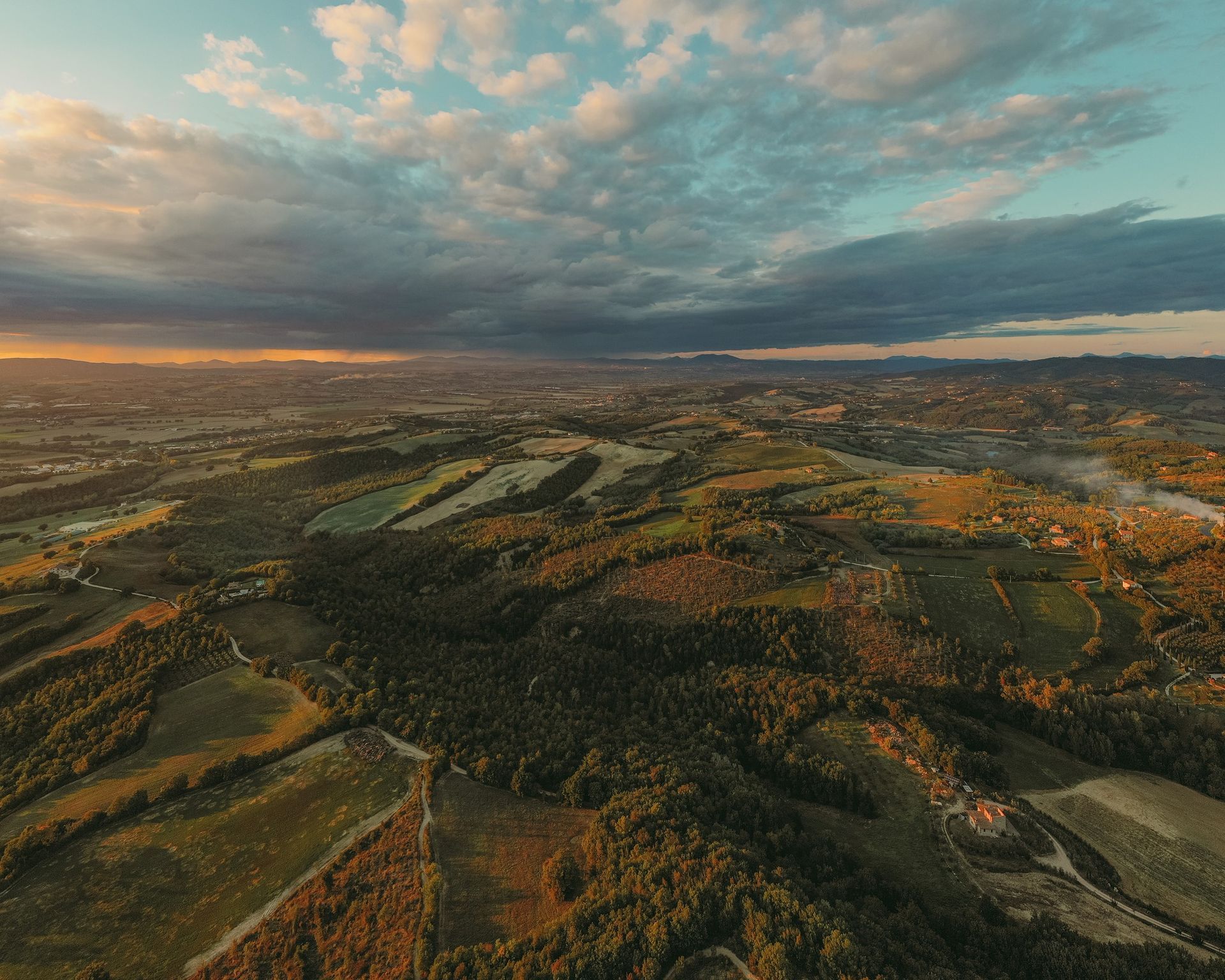 A wide landscape view of rolling hills, farmland, and vineyards surrounding Agri Segretum under a dramatic sky.