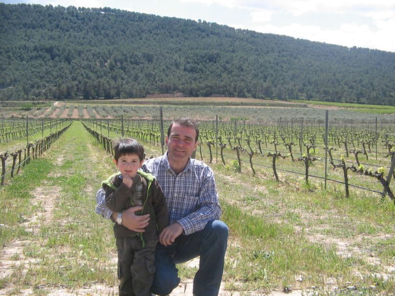 Man kneeling with a child in front of a vineyard, green grass, and trees in the background, at Pago Casa Gran.