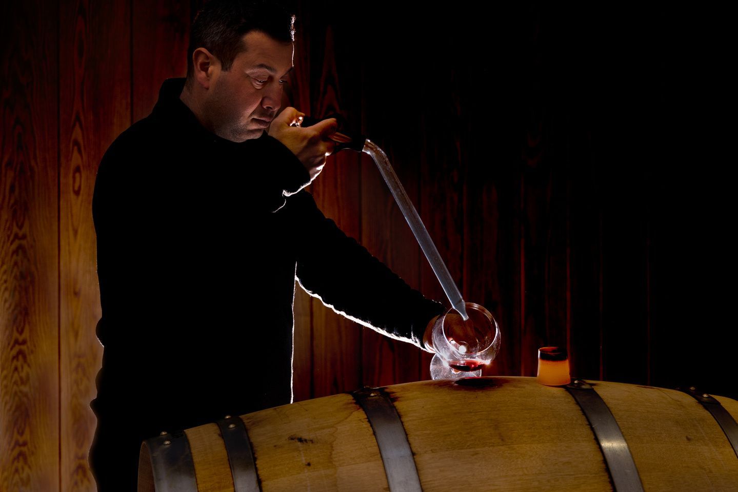 Alberto Voerzio using a wine thief to draw wine from a wooden barrel in a dark cellar.