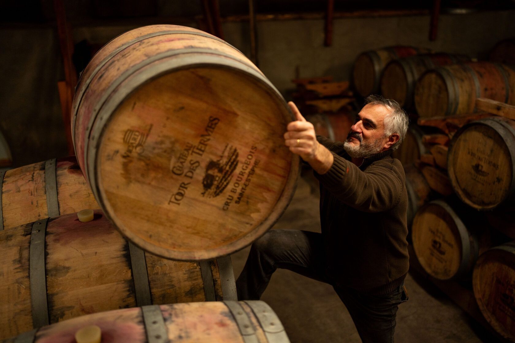 Man rolling a wooden wine barrel in a cellar, at Château Tour des Termes.