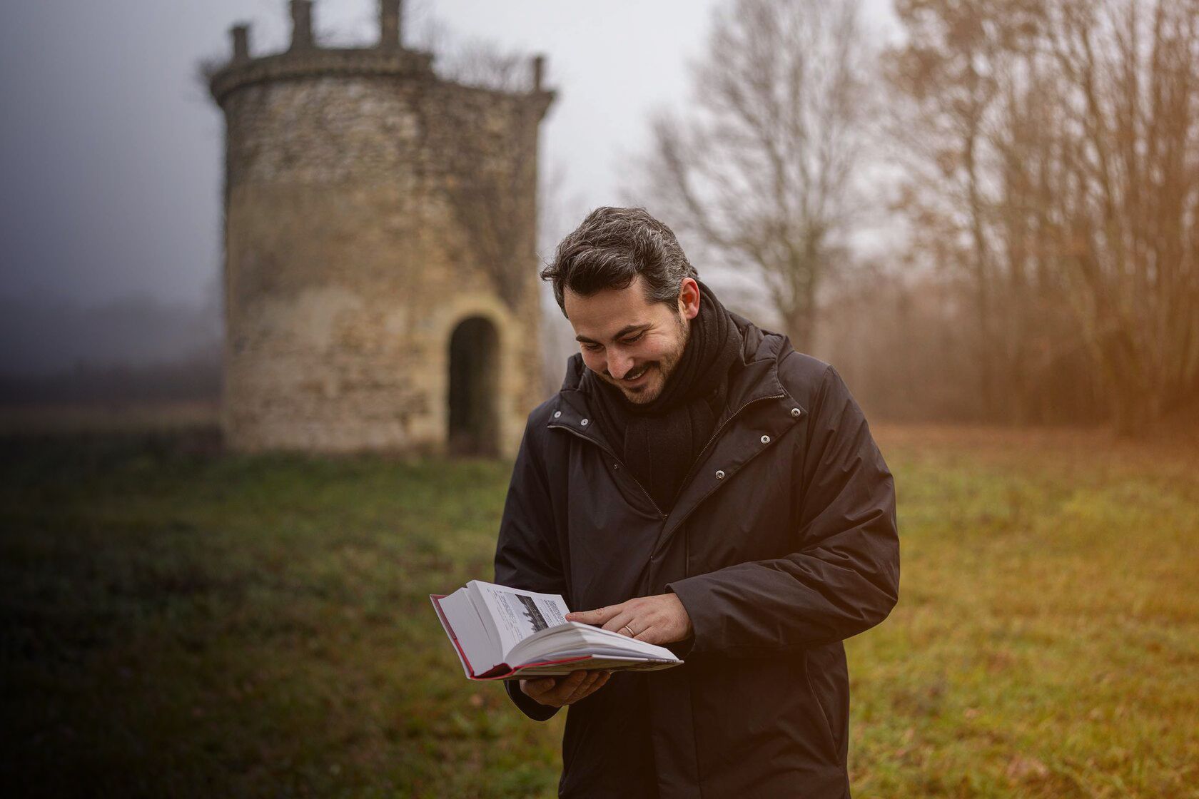 Man reading a book outdoors near a stone tower, at Château Tour des Termes.