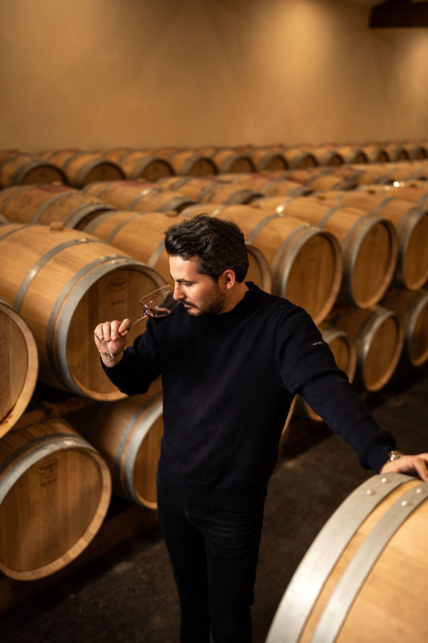 Winemaker of Château Tour des Termes tasting wine in a cellar lined with wooden barrels.