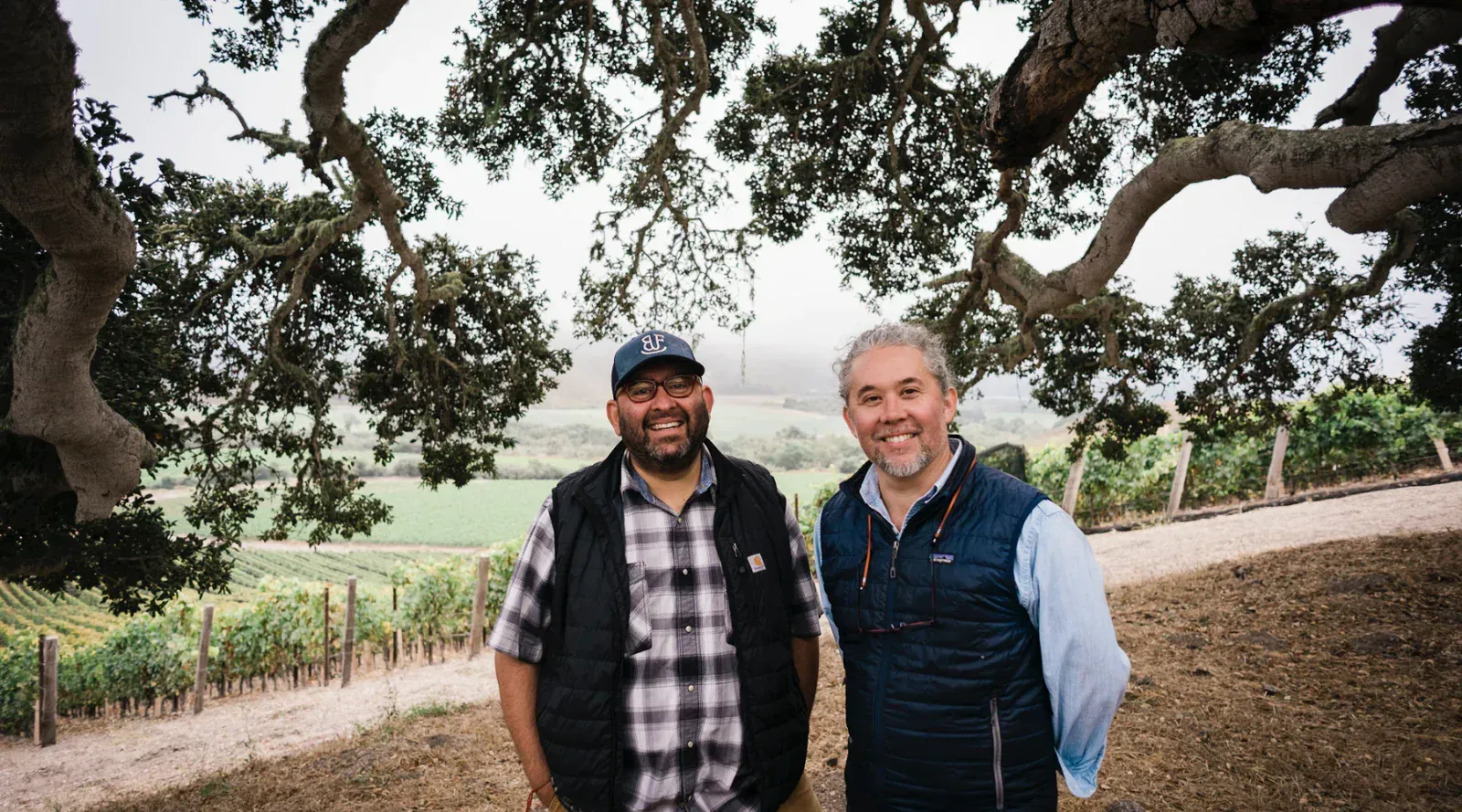 Two winemakers smiling in front of an Evening Land vineyard, with trees on the background.