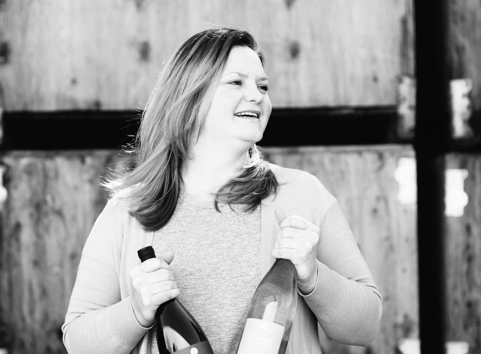 Woman holding two wine bottles, with wooden barrel background.