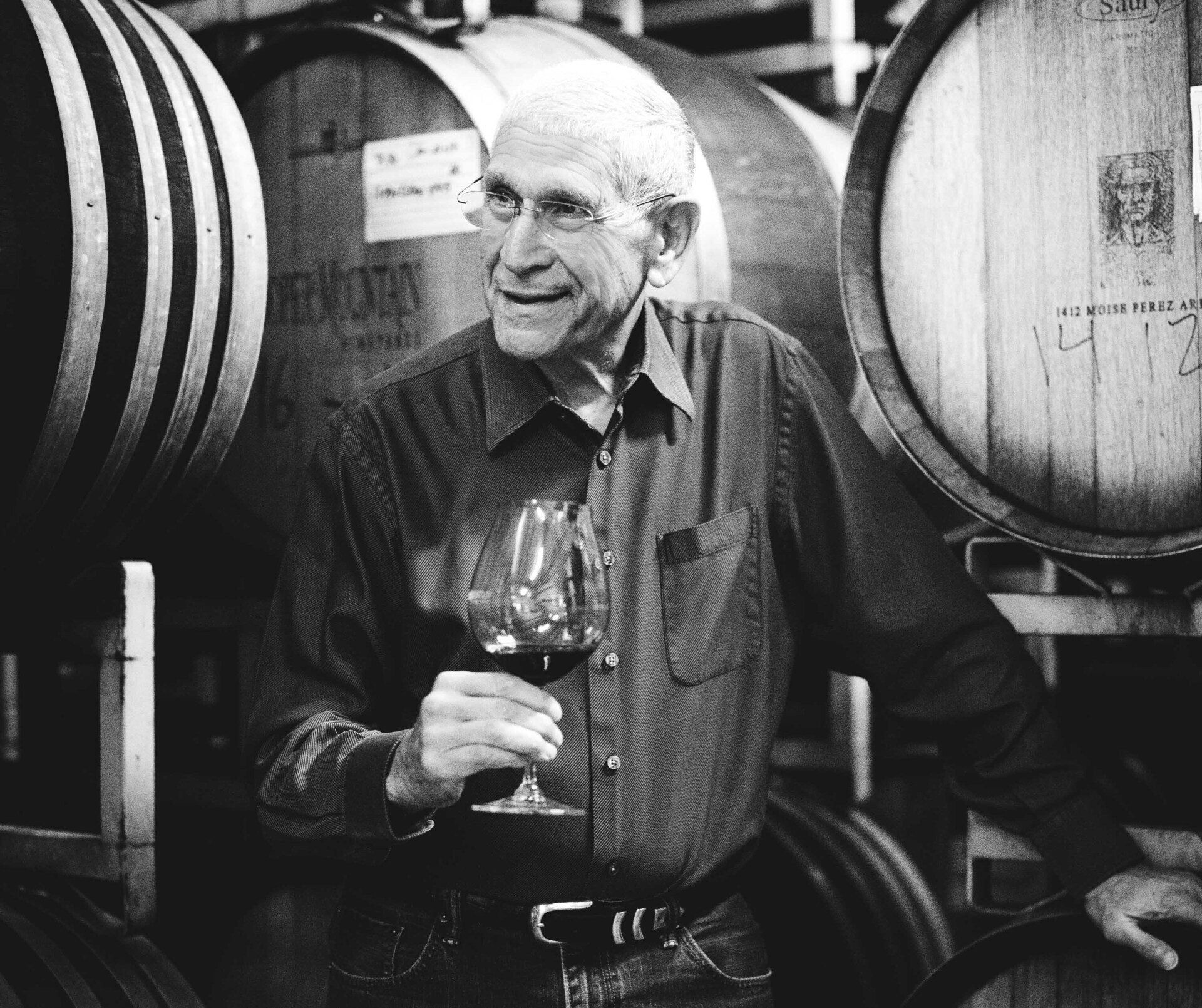 Man in a wine cellar holding a glass of red wine, surrounded by wooden barrels.