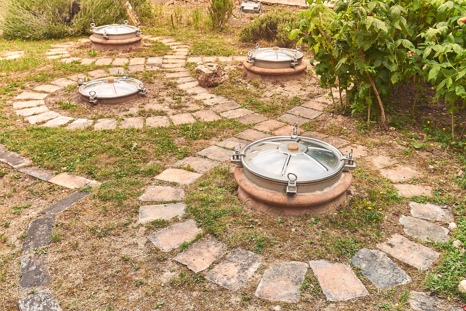 Braunstein - Circular lids on burried amphorae, surrounded by stone pathways and grass in an outdoor setting.