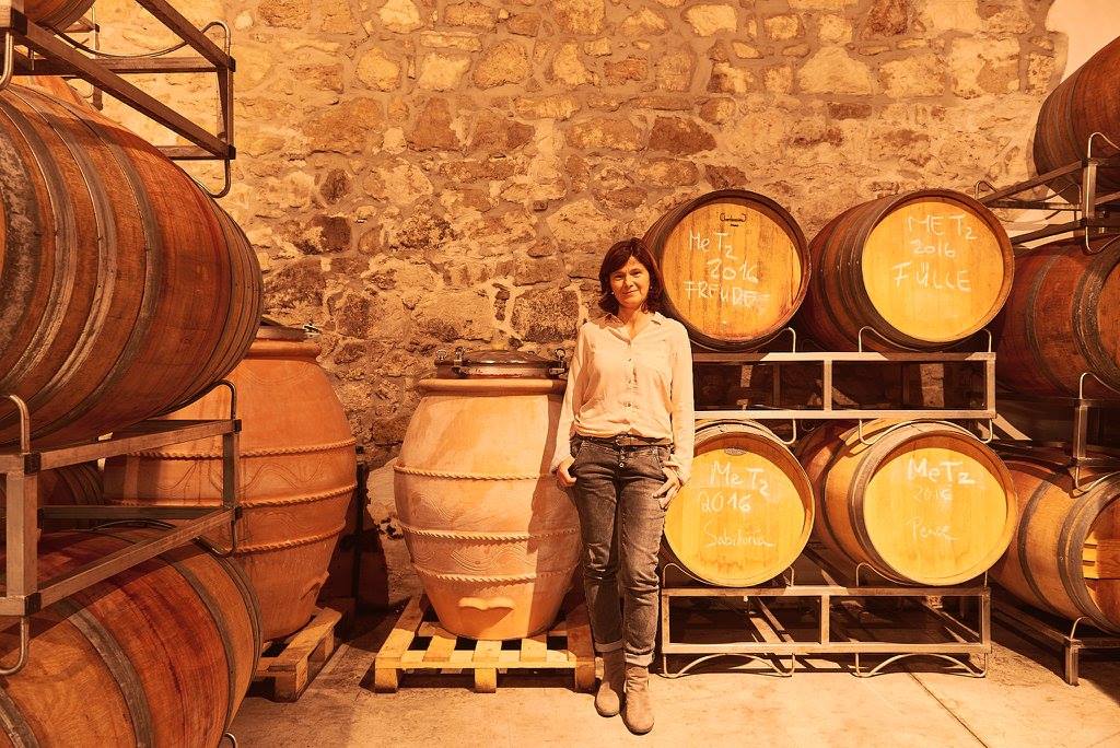 Birgit Braunstein standing in a wine cellar with barrels and a large clay pot.