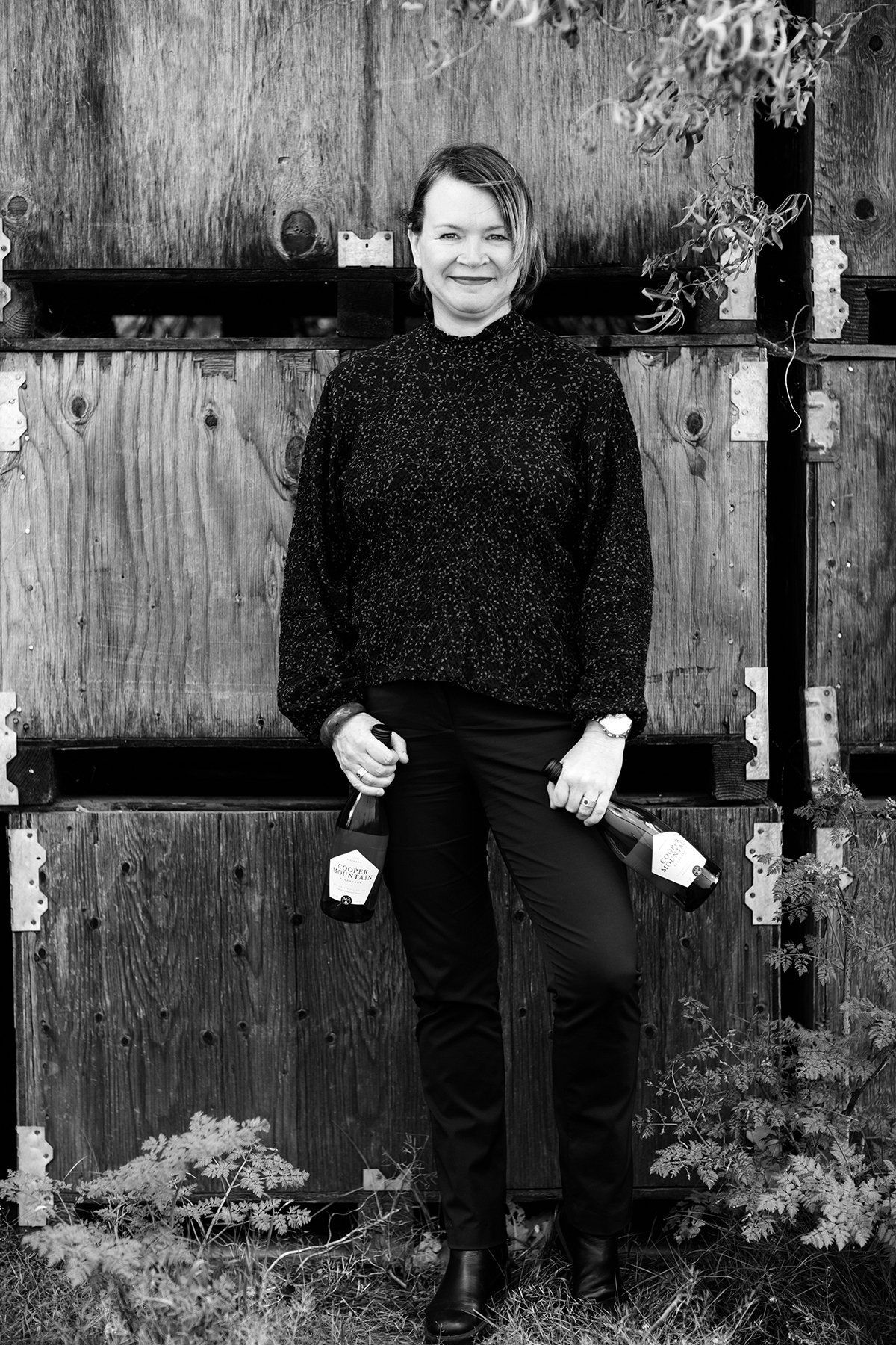 Black and white photo of woman holding wine bottles, standing in front of wooden crates.