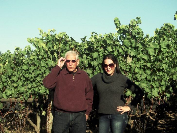 Man and woman wearing sunglasses stand in front of grapevines on a sunny day.