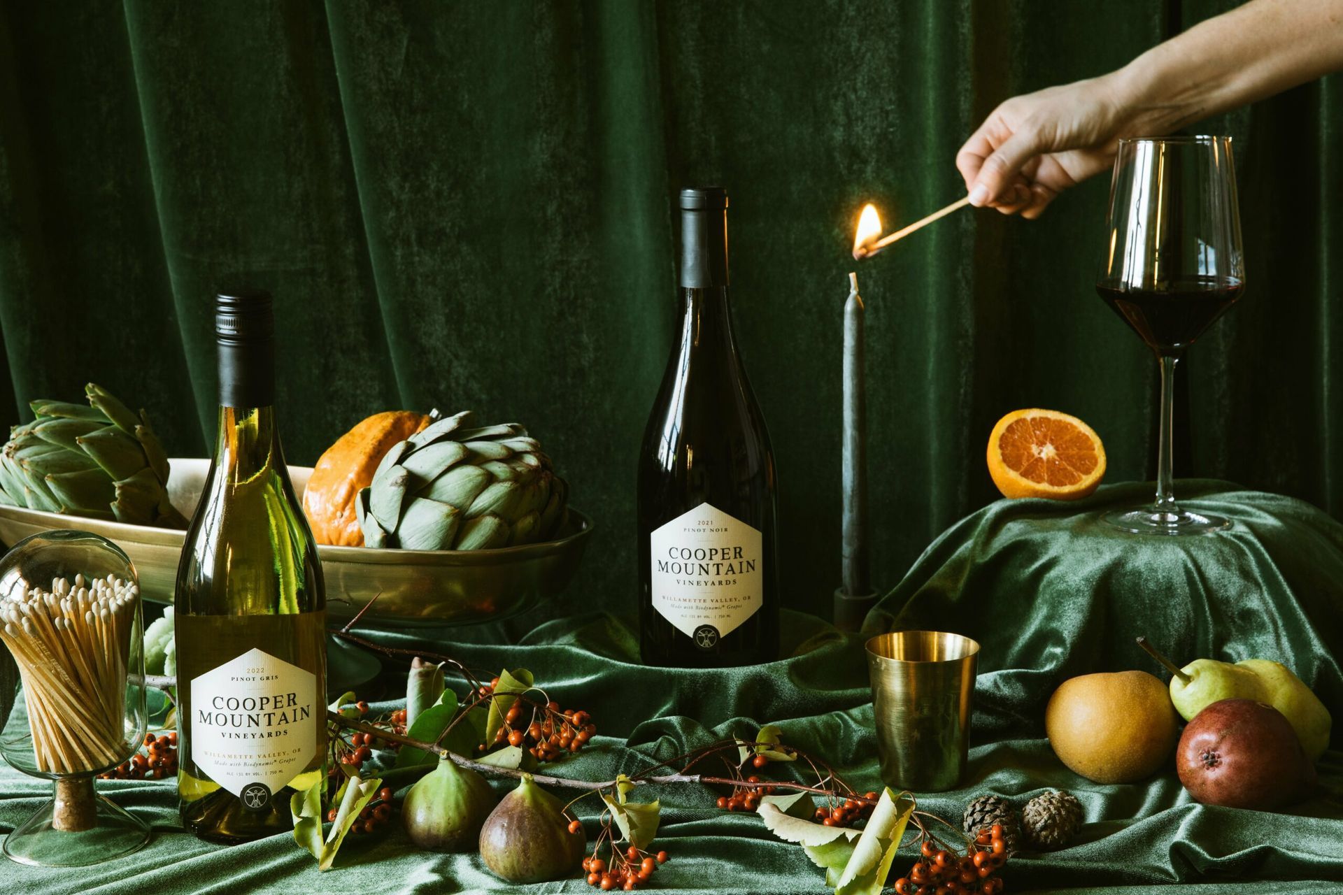 Image of Cooper Mountain wine bottles, with fruit, and candle being lit on green velvet.
