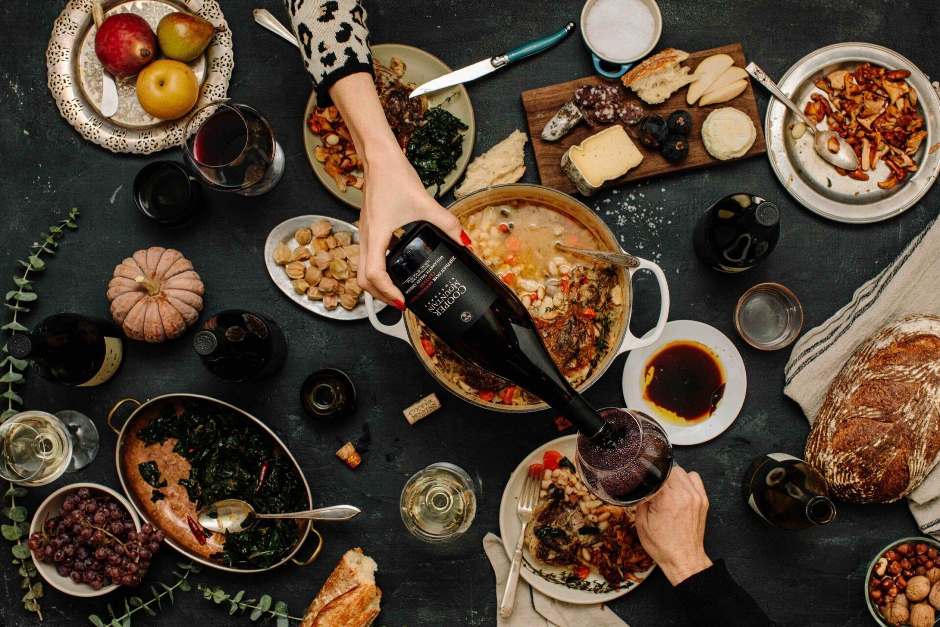 Overhead shot of a table with food, wine glasses, and a person pouring red wine.