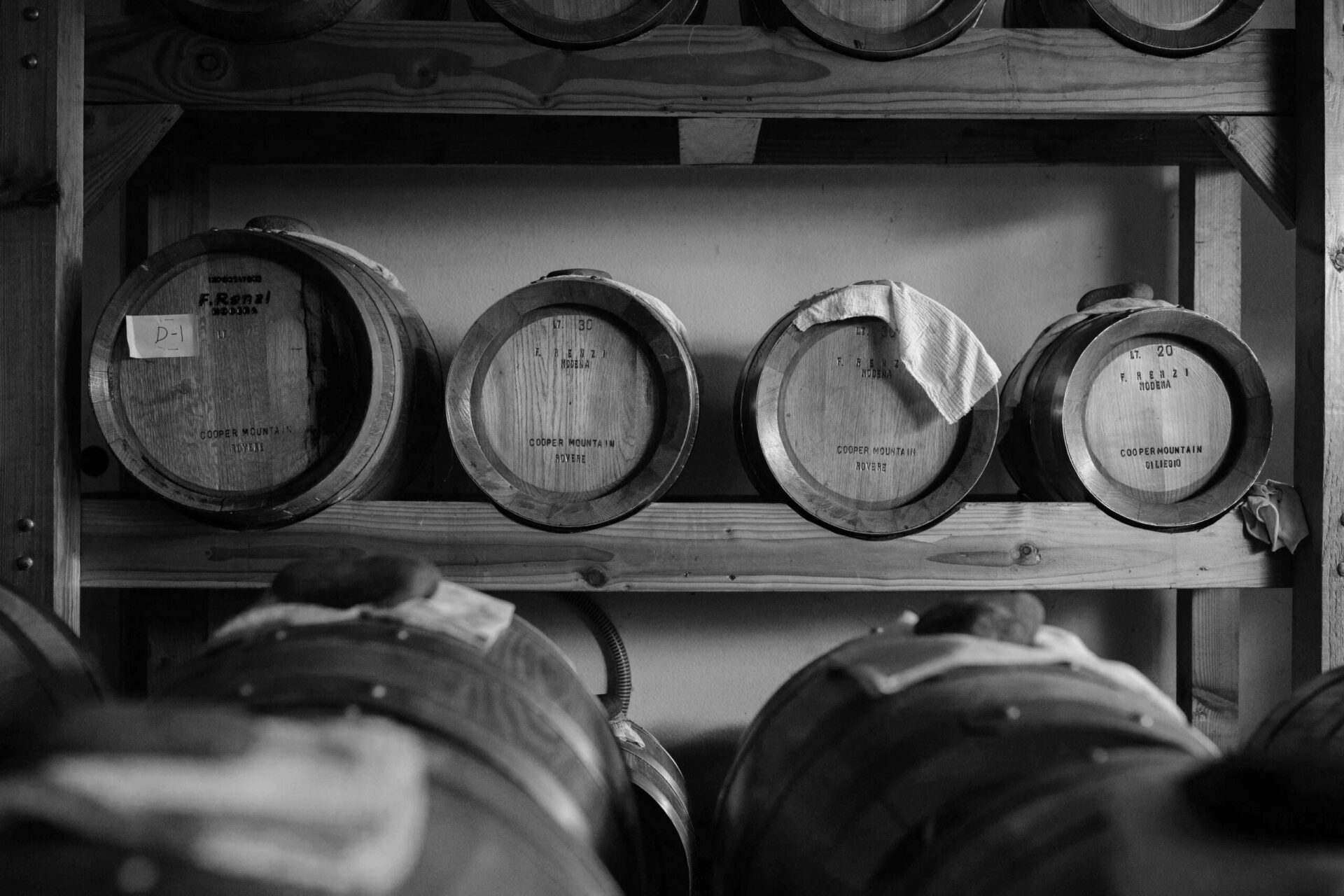 Wooden barrels on shelves in a cellar, some with cloth coverings.
