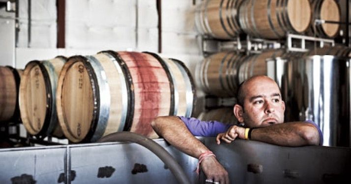 Man leans on a metal tank in Sandhi winery, surrounded by wooden barrels.