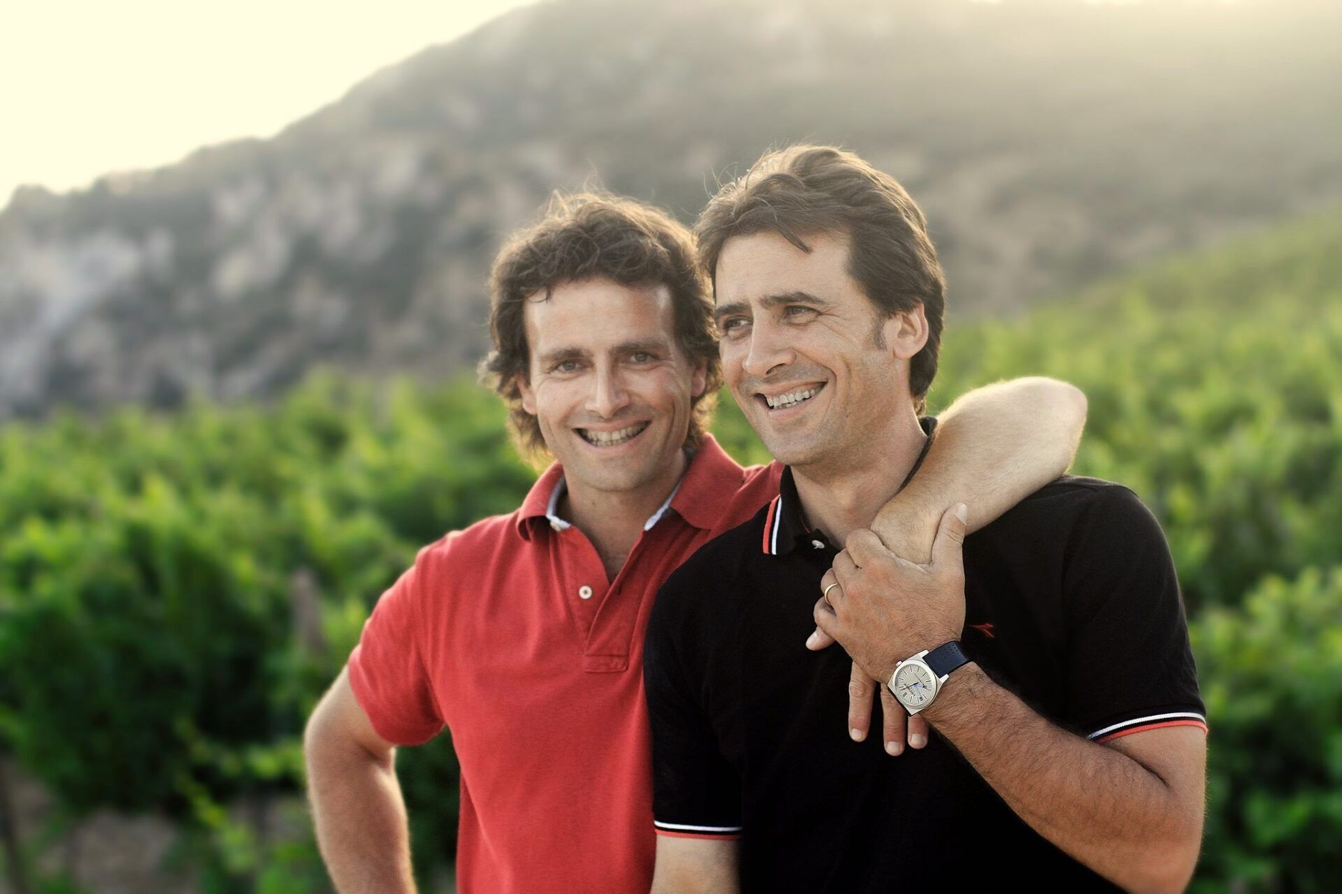 Di Giovanna winemakers with arms around each other, smiling, against a vineyard and mountain backdrop.