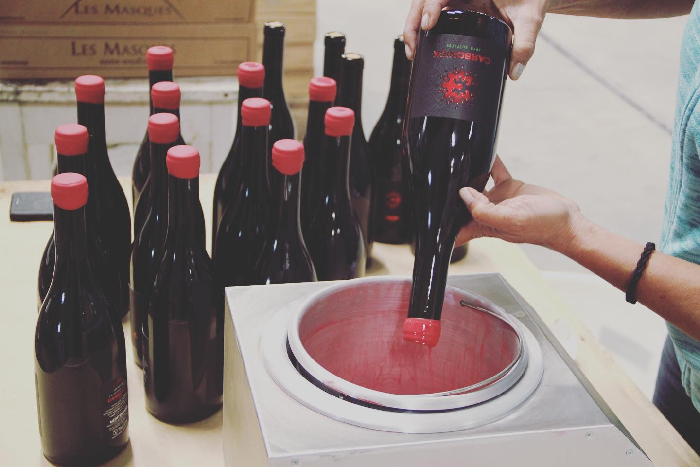 Person dipping a wine bottle neck into red wax, surrounded by other wine bottles, at Domaine des Masques winery.