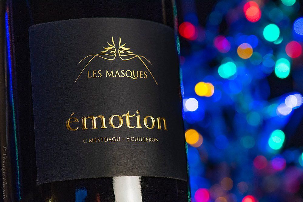 Close-up of a Domaine des Masques black wine bottle label with gold lettering, in front of colorful lights.