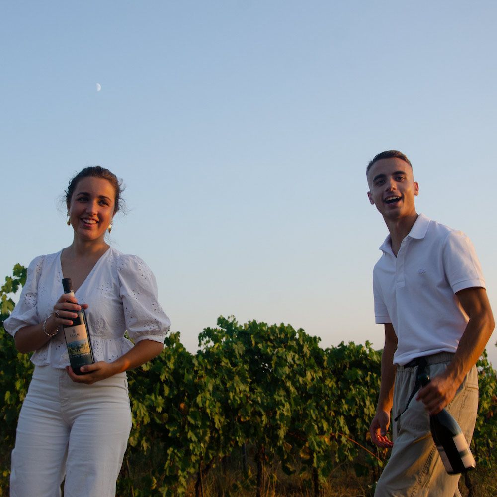 Two people in white attire holding wine bottles in a vineyard, smiling at the camera.
