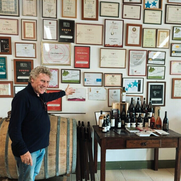 Paolo Controneo points at certificates on a wall, standing near wine bottles and a barrel.