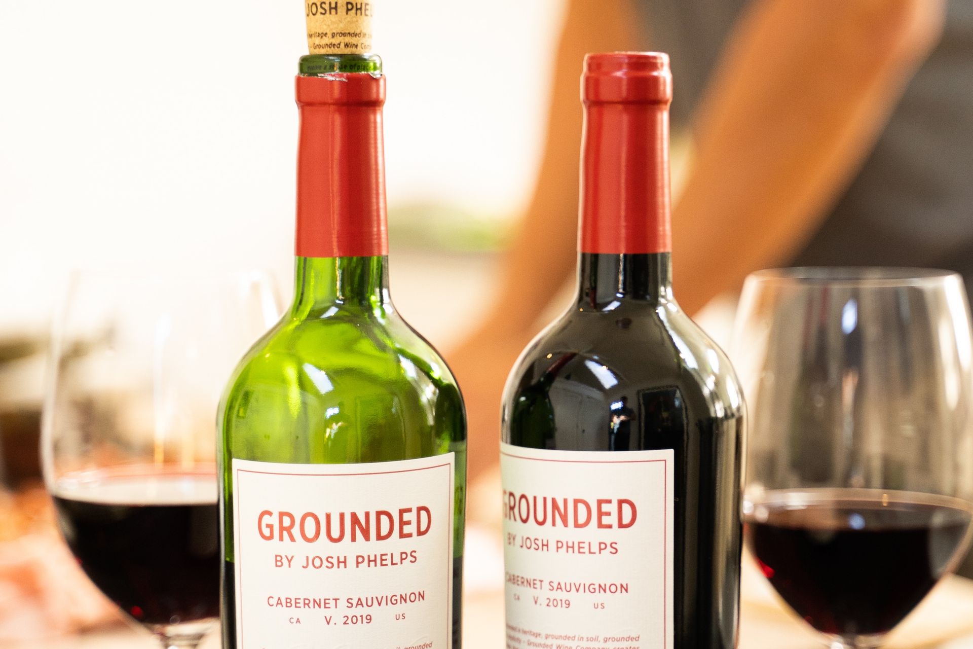 Two red wines of Grounded Wine Co, with glasses on the background.